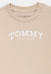 Κρεμ μπλουζάκι από βαμβάκι με στρογγυλή λαιμόκοψη, με τις λέξεις "TOMMY" και "HILFIGER" σε υψωμένα λευκά γράμματα. Μικρό κουμπί κοντά στην κολάρο.