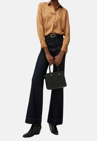 Camicia in satin marrone, jeans a vita alta blu scuro a gamba larga, borsa nera con doppi manici e stivali alla caviglia con zeppa. Design e texture puliti.