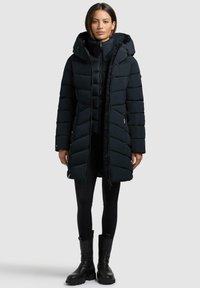 Schwarze Pufferjacke mit Kapuze, mit Reißverschluss vorne, horizontaler Steppung und seitlichen Taschen, getragen über schwarzen Leggings und Stiefeln.