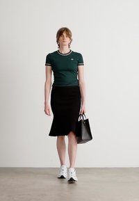 Tricou verde ajustat cu accente albe și negre, fustă neagră evazată, adidași albi și o geantă mică neagră. Fundal simplu.