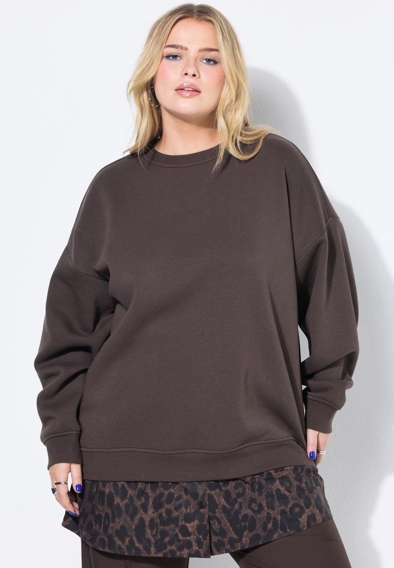 Sudadera marrón de gran tamaño con hombros caídos y cuello acanalado, superpuesta sobre una camisa de estampado de leopardo. Textura suave, diseño casual.