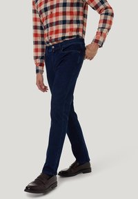 Ein Mann trägt dunkelblaue Jeans, braune Leder-Loafer und ein kariertes Hemd in Rot, Beige und Marineblau, während er seine Tasche richtet.