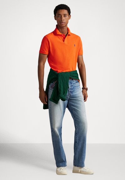 Polo orange, pull vert noué autour de la taille, jean bleu clair, baskets beige. Coupe droite, col classique, marque discrète visible.