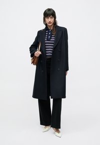 Lange marineblauwe overjas met dubbelzijdig ontwerp. Daaronder een gestreept crop top en zwarte broek met wijde pijpen. Witte puntige platte schoenen en een bruine crossbodytas.