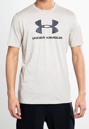 Beżowa koszulka Under Armour z krótkim rękawem, z czarnym logo i napisem, noszona przez osobę w czarnych spodniach na białym tle.