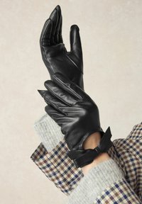 Schwarze Lederhandschuhe mit einer strukturierten Oberfläche; verfügen über spitze Finger und ein Bändchen am Handgelenk. Der Hintergrund zeigt graue Strick- und Karomuster.