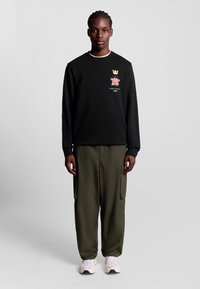 Schwarzer Sweatshirt mit Rundhalsausschnitt, versehen mit einem floralen Emblem. Kombiniert mit olivgrünen Cargo-Hosen und weißen Sneakers.