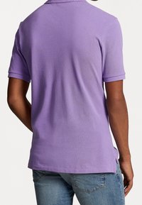 Man som bär en kortärmad lavendelfärgad polo och blå jeans, sedd bakifrån mot en enkel vit bakgrund.