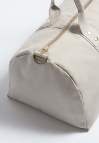 Beige canvas duffelbag met een ronde basis, gouden rits en metalen accenten. Bevat dubbele stoffen handvatten voor eenvoudig dragen.