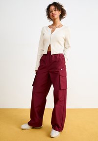 Rippiger, cremefarbener cropped Cardigan mit einem Reißverschluss vorne, kombiniert mit lockeren, bordeauxroten Cargo-Hosen mit Seitentaschen und einem weißen Logodetail.