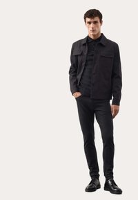 Czarna kurtka z dwoma przednimi kieszeniami, noszona na czarnym polo i dopasowanych czarnych dżinsach. Obuwie to eleganckie czarne buty, tworzące monochromatyczny look.