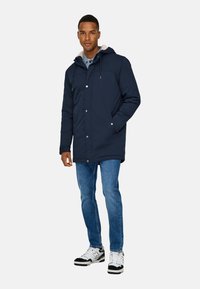 Marinblå parkas med mjukt foder i huvan, knappstängning och sidofickor, i kombination med ljusblå jeans och svart-vita sneakers.