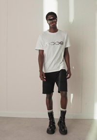 Λευκό t-shirt με ανακλαστικό γραφικό, συνδυασμένο με μαύρα σορτς και μαύρες δερμάτινες μπότες με κορδόνια. Απαλά υλικά και απλός σχεδιασμός.