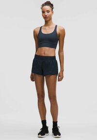 Tumšs sporta krūšturis un augsta vidukļa šorti, abi izgatavoti no viegla materiāla. Modelis valkā melnas sporta kurpes ar baltu zoli.