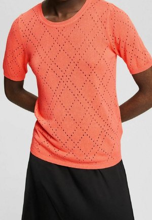 Osoba nosząca koralowy krótkim rękawem sweter z dzianiny z diamentowym wzorem ażurowym i czarną spódnicę na jednolitym tle.