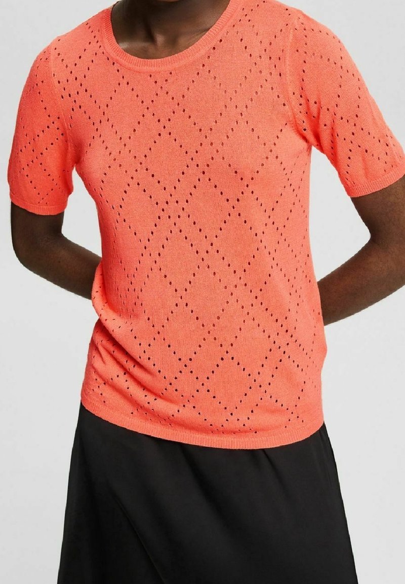 Personne portant un haut en maille corail à manches courtes avec un motif œillet en forme de losange et une jupe noire sur un fond uni.