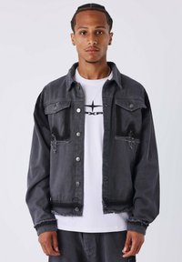 Giacca in denim nera con bordi sfilacciati, due tasche sul petto e bottoni argentati. Indossata sopra una t-shirt bianca con un design grafico nero.