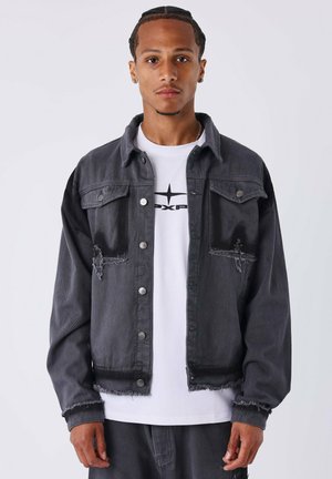 Zwarte denimjack met rafelige randen, twee borstzakken en zilveren knopen. Gedragen over een wit T-shirt met een zwart grafisch ontwerp.