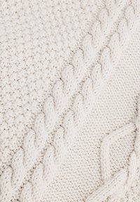 Krémszínű kötött anyag, amely texturált mintázattal rendelkezik, emelt, fonott részletekkel átszőve. Az összkép egységes és koherens.