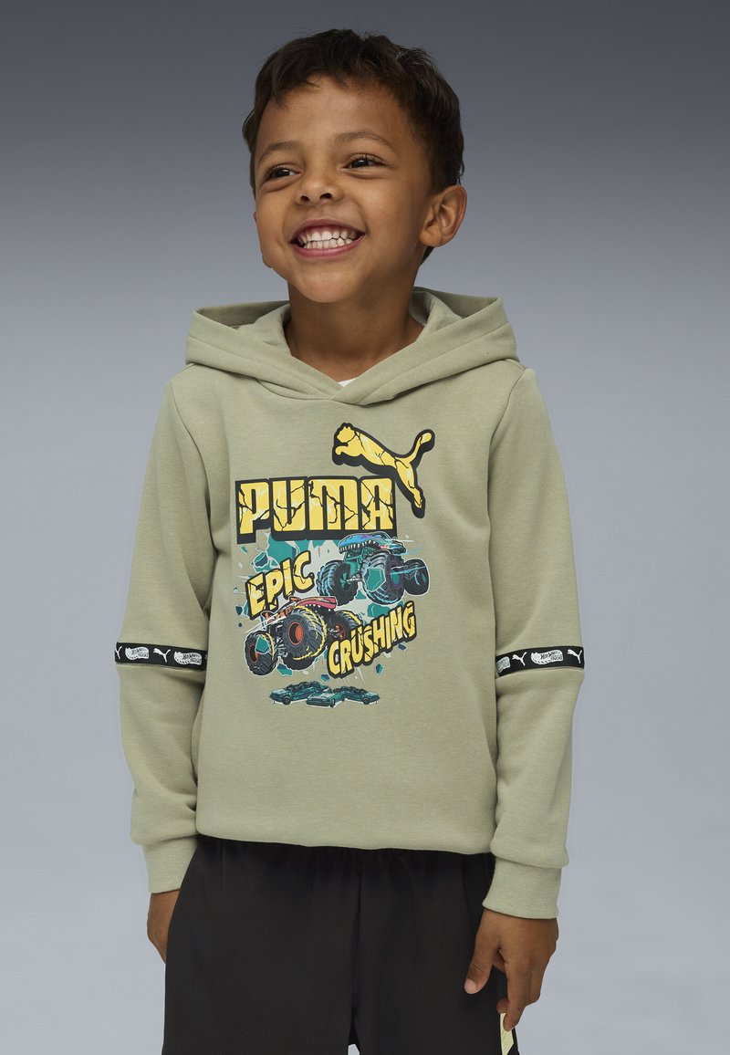 Ragazzo sorridente che indossa una felpa Puma verde chiaro con grafica di camion mostro e testo "Epic Crushing", abbinata a pantaloni neri, in piedi contro uno sfondo grigio.