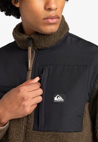 Fleece jacket with black quilted panels and olive green textured fabric. Features a zippered pocket and logo patch on the chest.