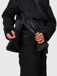 Manos ajustando el zipper frontal y las capas interiores de una chaqueta impermeable negra sobre pantalones cargo negros con bolsillos.