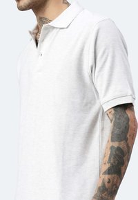 Hellgraues Poloshirt mit weicher Textur, dreiknöpfiger Knopfleiste und kurzen Ärmeln. Vorgestellt an einem Modell mit Tattoos am Arm.