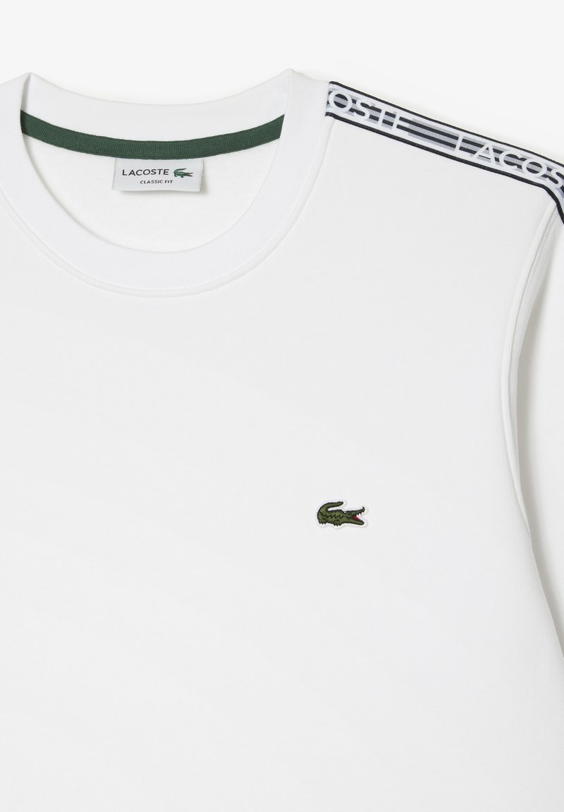 T-shirt en coton blanc avec col vert, logo crocodile vert brodé sur la gauche et accents d'épaule à rayures noires. Design à coupe classique.