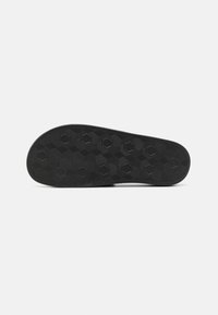 Semelle de chaussure noire avec motif de bande de roulement géométrique hexagonal et texturé sur fond blanc uni.