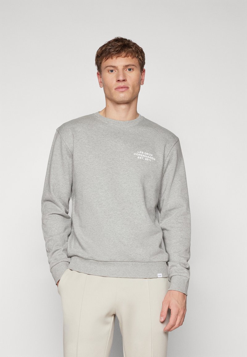 Uomo che indossa un maglione grigio chiaro con la scritta "LES DEUX COPENHAGEN EST. 2011" sul petto e pantaloni beige, in piedi contro uno sfondo neutro.