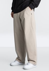 Beige vide bukser laget av bomull, med sidelommer. Kombinert med hvite joggesko, som viser en avslappet, uformell design.