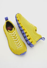 Κίτρινα πλεκτά αθλητικά παπούτσια με μωβ λεπτομέρειες, που διαθέτουν ανατομική σόλα, σχέδιο με κορδόνια και υφή για καλύτερη αναπνοή.