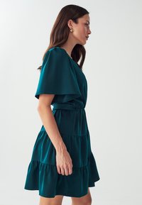 Teal-blaues, knielanges Kleid aus einem weichen Stoff, mit einem ausgestellten Ärmeldesign, taillierter Taille und geschichteten Rüschendetails.