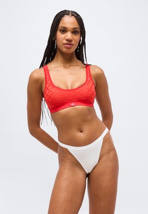Γυναίκα με μακριά κοτσίδες που φοράει κόκκινο αθλητικό σουτιέν με σχέδια και λευκά εσώρουχα με ψηλή γραμμή, στέκεται με τα χέρια πίσω από την πλάτη της.