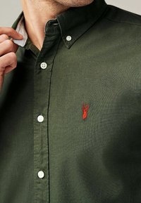 Ciemnozielona koszula zapinana na guziki, wykonana z fakturowanej tkaniny, z białą listwą guzikową i czerwoną haftowaną grafiką jelenia na piersi.