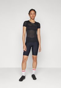 Svart mesh kortermet jersey med logo, kombinert med mørke sykkelshorts med røde detaljer. Modellen har på seg hvite sokker med røde striper.