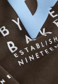 Brun t-shirt med store blå og hvide bogstaver, glat bomuldsstruktur og afslappet pasform.