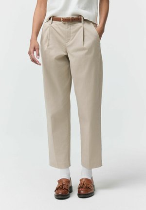 Kobieta ubrana w beżowe spodnie o prostych nogawkach z plisami z przodu, brązowy skórzany pasek, białe skarpetki i brązowe mokasyny z chwostami, stojąca z jedną ręką w kieszeni.