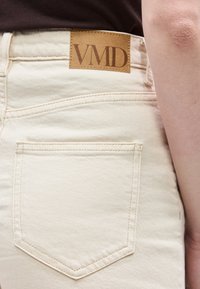 Κοντινό πλάνο της πίσω τσέπης σε μπεζ τζιν και δερμάτινο λογότυπο με την επιγραφή «VMD», φορεμένα με σκούρο τοπ, που δείχνει μέρος του βραχίονα ενός ατόμου.