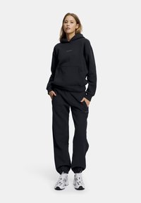 Svart bomull hettegenser med frontlomme og logo. Matchet med svarte joggebukser, med sidelommer og tettsittende mansjetter. Brukt med sølvfargede sneakers.