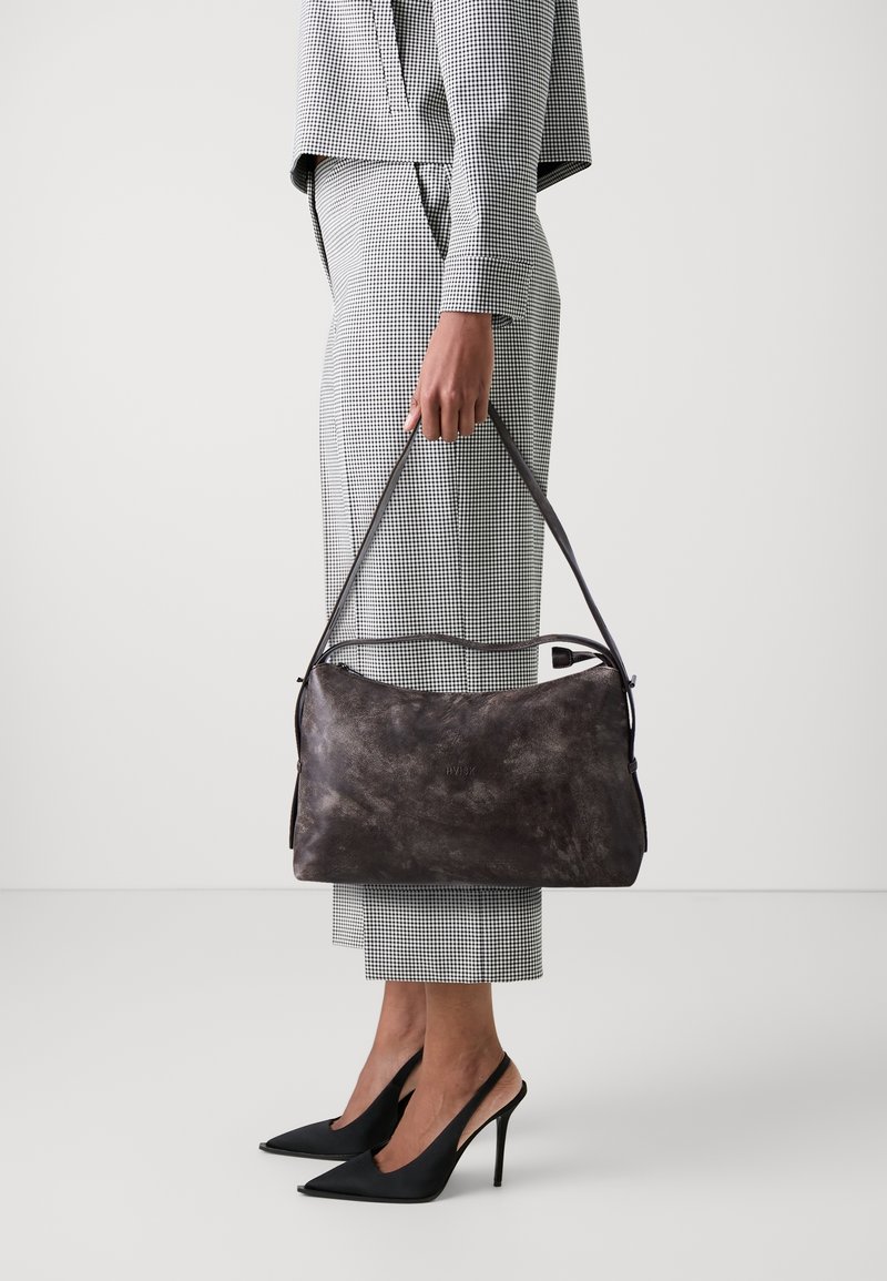 Personne en costume à carreaux tenant un grand sac à main en cuir foncé, portant des talons hauts pointus noirs, debout devant un fond blanc uni.