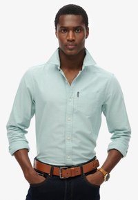 Camicia azzurra chiara con colletto e taschino sul petto, abbinata a una cintura marrone. Il modello indossa jeans scuri e un orologio d'oro.