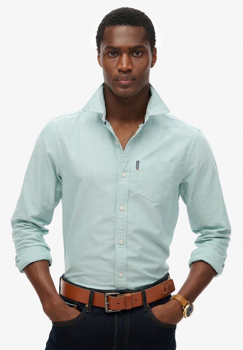 Camicia azzurra chiara con colletto e taschino sul petto, abbinata a una cintura marrone. Il modello indossa jeans scuri e un orologio d'oro.