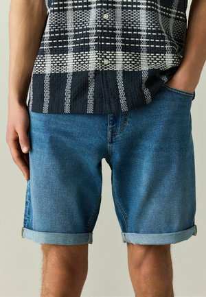 Férfi, aki kék, felhajtott szárú farmer rövidnadrágot és fekete-fehér mintás rövid ujjú inget visel, kezével a zsebében, áll egy egyszínű háttér előtt.