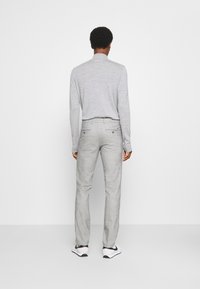 Man med ljusgrå turtleneck-tröja, grå skräddarsydda byxor och vita Nike-sneakers, stående vänd mot en vit vägg.