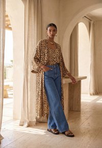 Žena s uhlazenými vlasy, oblečená v topu s leopardím vzorem a dlouhém odpovídajícím kimonu, modré široké džíny, stojící u kamenného stolu uvnitř budovy.