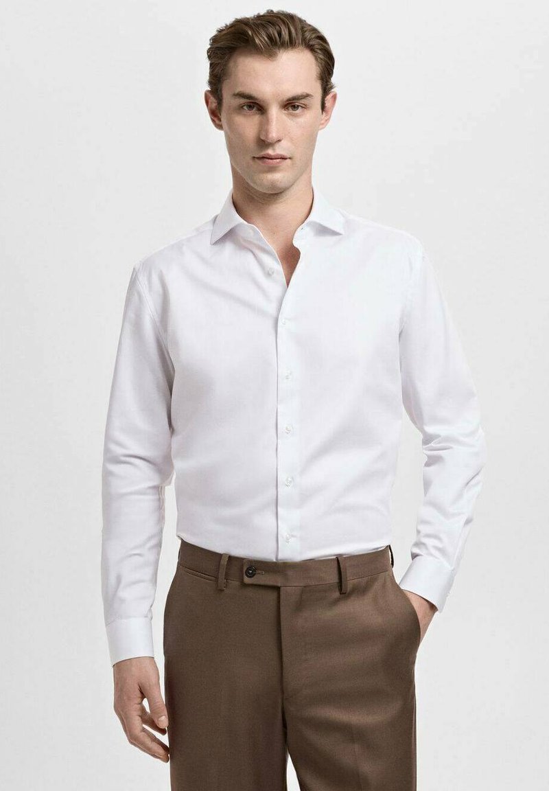 Cămașă albă cu mâneci lungi, guler îngust și nasturi pe față, asortată cu pantaloni maro eleganți. Material neted din bumbac.