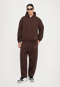 Hanorac maro supradimensionat cu buzunar frontal, combinat cu pantaloni de trening asortați. Accesoriile includ ochelari de soare negri și adidași albi.