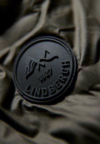 Svart gummimärke med "LINDBERGH 1927" på en strukturerad, olivgrön tygbakgrund, med mjukt, skrynkligt material.