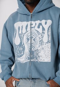 Blauer Kapuzenpullover mit Reißverschluss und großem weißem Grafikdesign, das ein Gesicht und abstrakte Muster zeigt. Hergestellt aus weichem, strukturiertem Stoff.